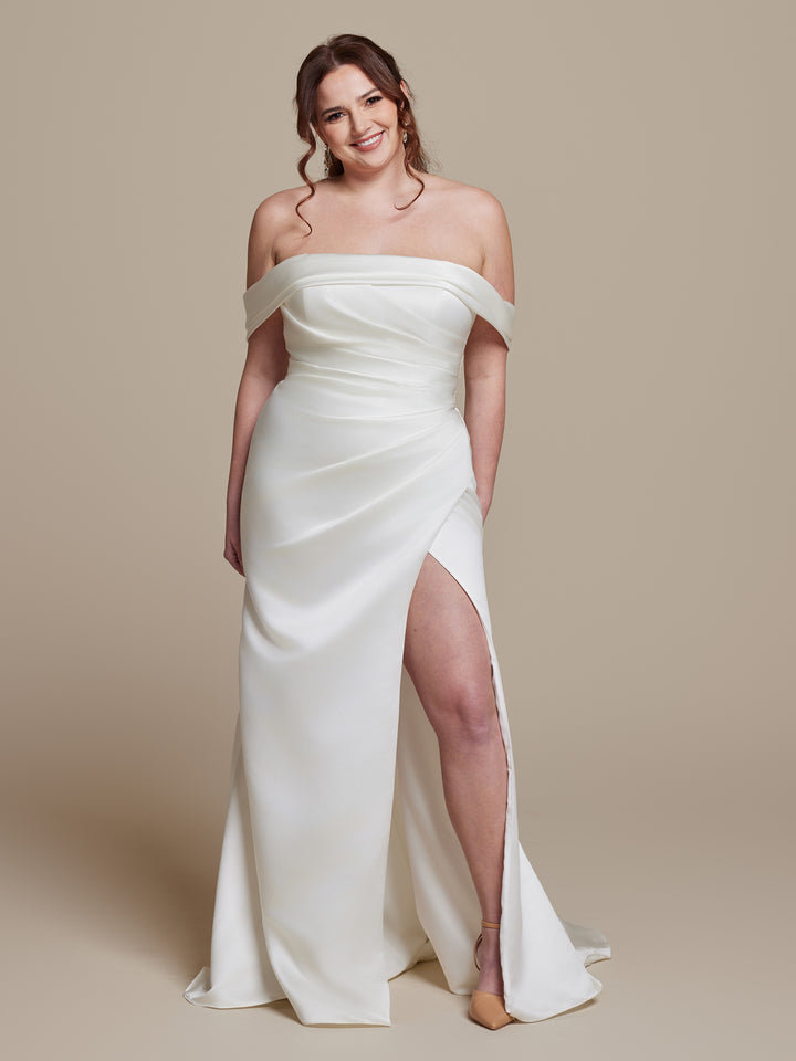 A woman models the "Phoebe" off-the-shoulder wedding dress by averyaustin, featuring a thigh-high slit. Against a beige backdrop, her wavy brown hair is in a loose updo, paired with nude heels for an elegant look.