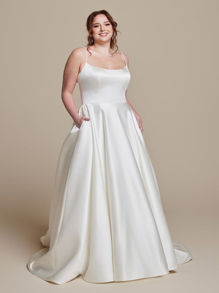 A woman smiles in the Millie dress by averyaustin—a simple ivory satin wedding gown with thin straps, pockets, and a fitted bodice. Its full A-line skirt flows to the floor, creating an elegant look against a plain beige background.
