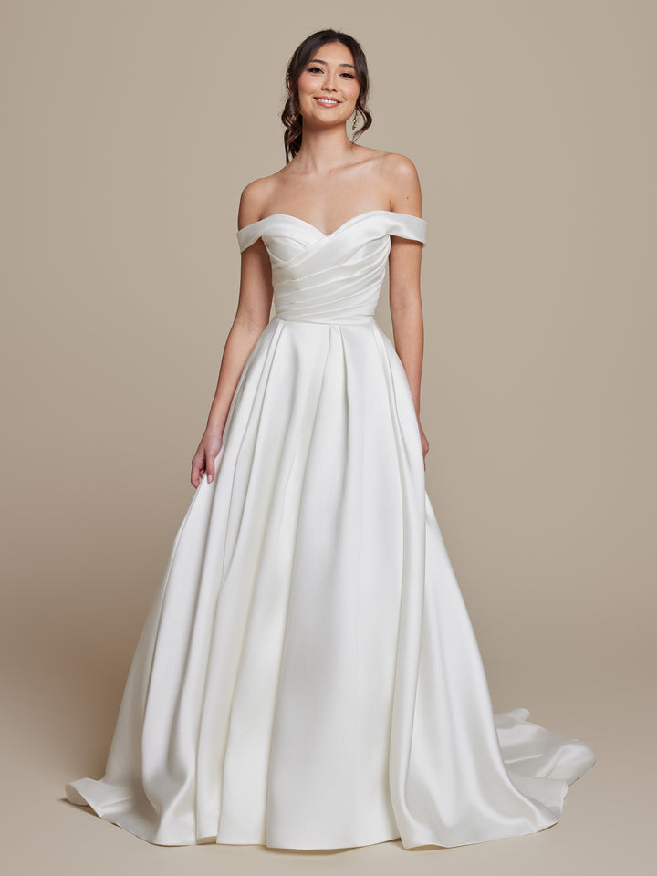 A woman smiles while wearing the "Frankie" ivory off-the-shoulder mikado wedding dress by averyaustin, featuring a fitted, ruched bodice and a full skirt, standing against a plain beige background.