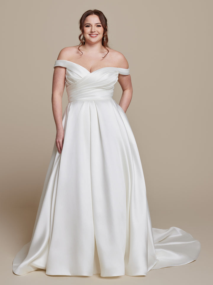 A woman smiles in the averyaustin "Frankie" ivory wedding dress, featuring an off-the-shoulder fitted bodice and full floor-length skirt. A plain beige background highlights the timeless elegance of her gown.