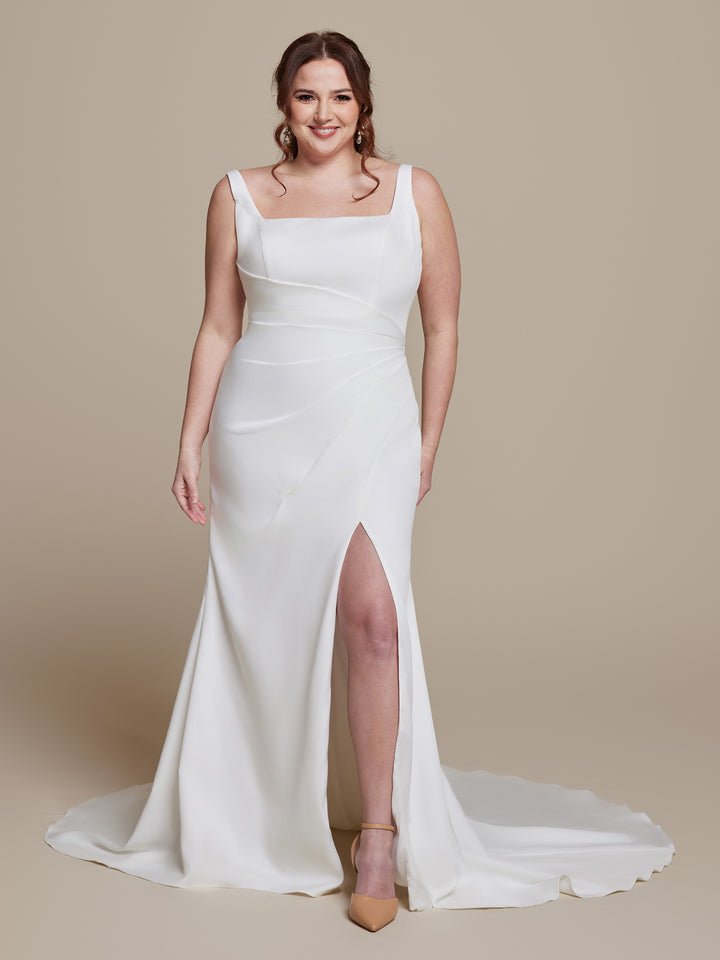 A woman smiles in the Darcey wedding dress by averyaustin, featuring ivory crepe fabric, a square neckline, and a thigh-high slit. She stands against a beige background in tan heels with her hair styled in loose waves.