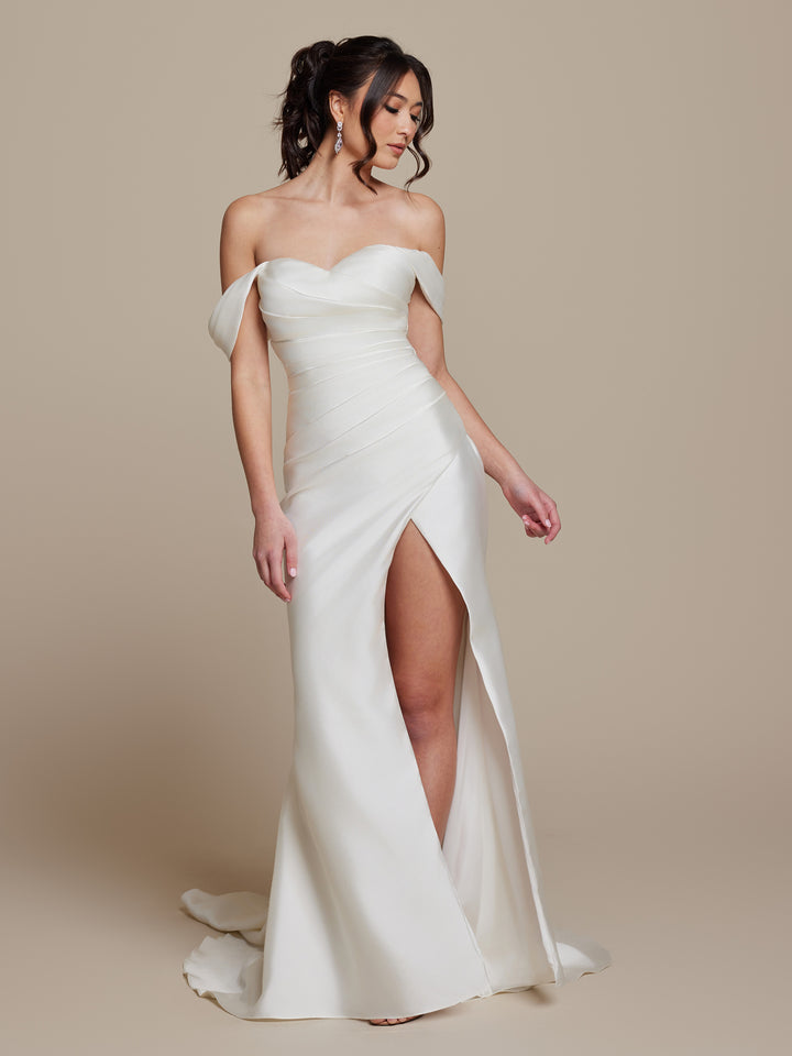 A woman in the Elodie by averyaustin, an ivory off-the-shoulder wedding dress with a sweetheart neckline and high thigh slit, poses against a neutral background.