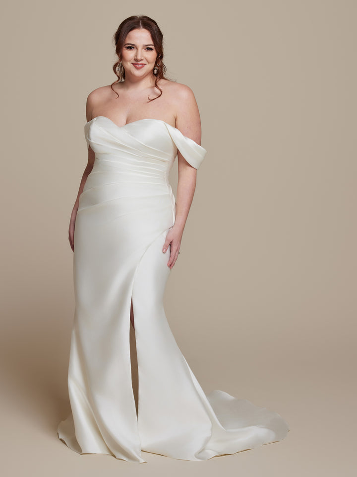 A woman wearing the Elodie ivory off-the-shoulder floor-length wedding dress by averyaustin, featuring a sweetheart neckline and front slit, stands against a plain beige background, smiling at the camera.