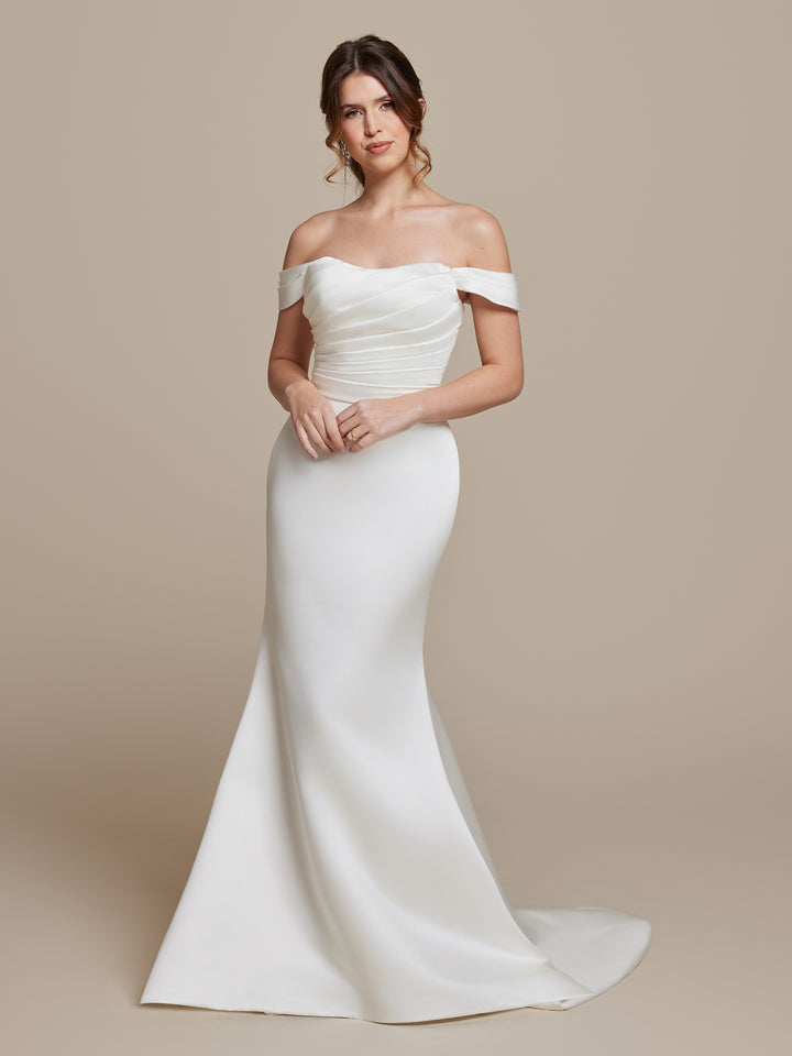 A woman in the Raya ivory off-the-shoulder fit-and-flare wedding dress by averyaustin stands against a plain beige background, looking at the camera with her hands gently clasped and a slight train flowing behind her.