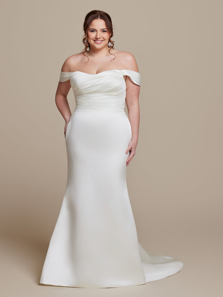 A woman in the averyaustin Raya wedding dress—an off-the-shoulder, ivory satin gown with a slight train—stands smiling against a neutral background, her brown hair styled in loose curls.