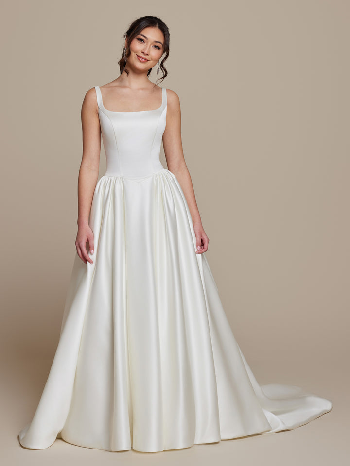 A woman wears the Tilly by averyaustin, an ivory satin wedding dress featuring a fitted bodice, square neckline, and full floor-length A-line skirt with a small train, set against a neutral background.