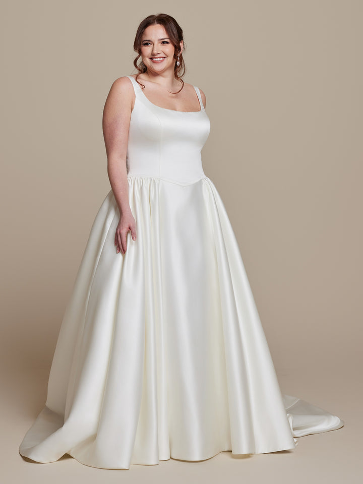 A smiling woman with long brown hair wears the averyaustin Tilly satin ball gown wedding dress in ivory with a square neckline, standing against a plain beige background.