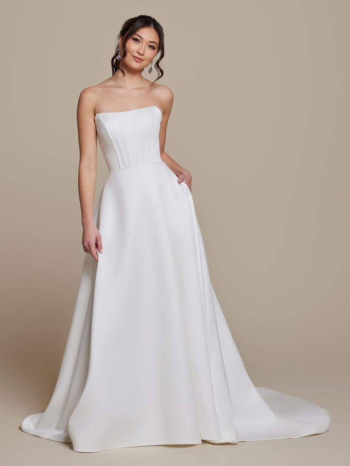 A woman in the Lyra strapless satin wedding dress by averyaustin, featuring a fitted bodice and flowing A-line skirt in ivory, stands against a plain beige background, smiling gently with one hand on her hip.