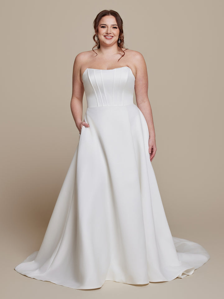 A woman wearing the Lyra strapless satin wedding dress by averyaustin in ivory, featuring a structured bodice and subtle pleats, stands against a neutral background, smiling.