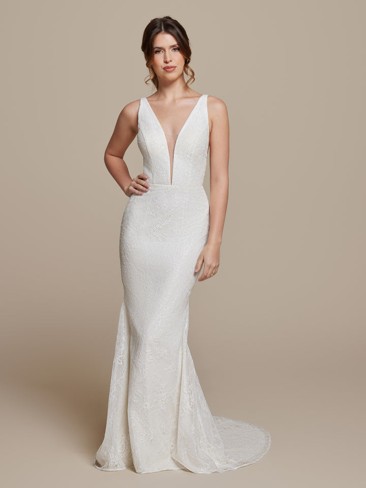 A woman stands against a plain beige background, wearing the averyaustin TH-Pippa wedding dress in Ivory/Nude/Nude—sleeveless with white lace, a deep V neckline, and a slight train.
