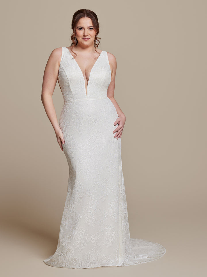 A woman models the averyaustin TH-Pippa wedding dress in Ivory/Nude/Nude, featuring a sleeveless lace design, deep V neckline, and fit-and-flare silhouette against a plain beige background.