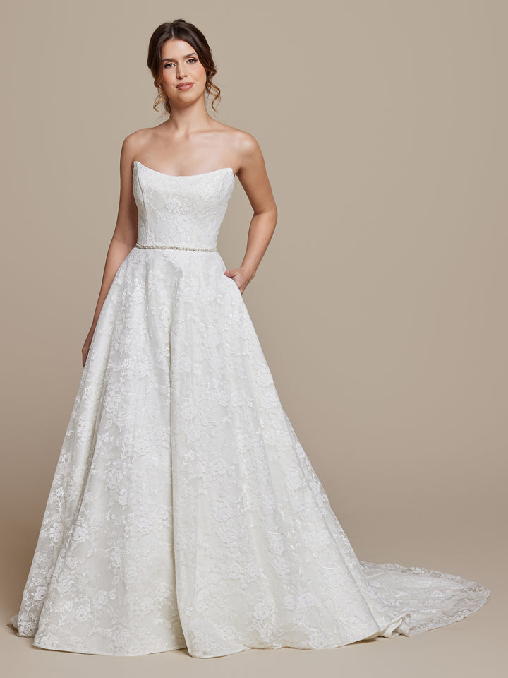 A woman stands against a plain background wearing the Freya wedding dress by averyaustin in Ivory Glitter, featuring a strapless fitted bodice, full skirt with short train, and pockets. Her hair is styled in loose waves.