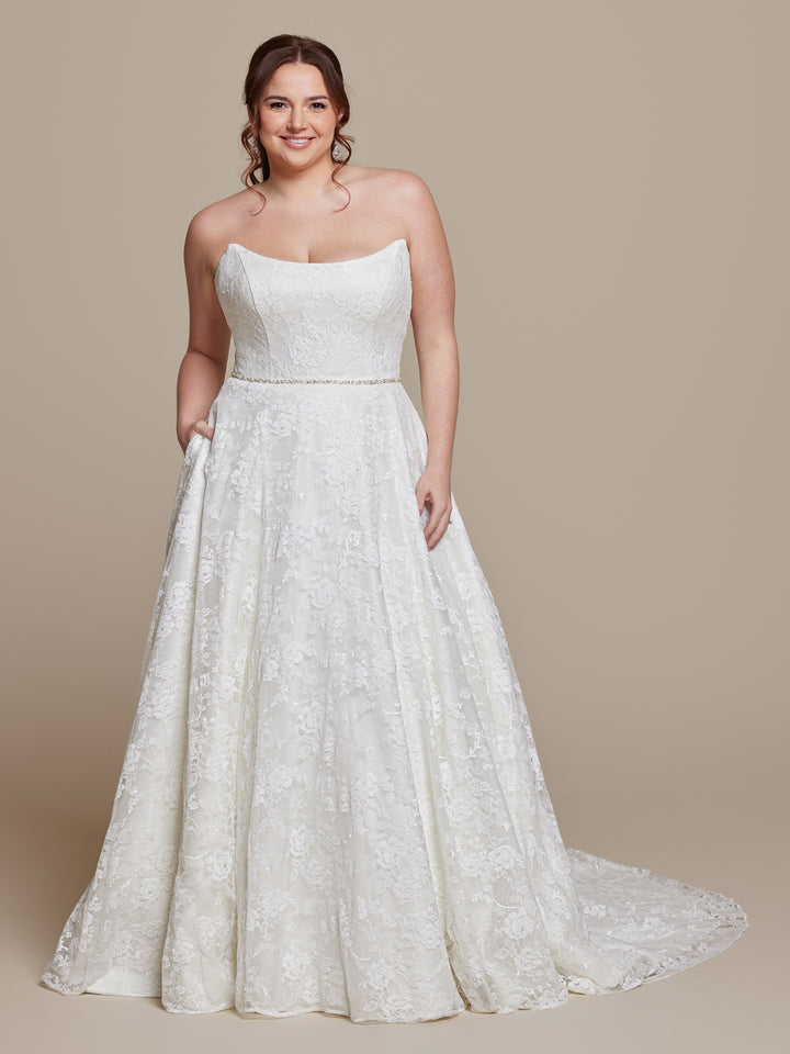A smiling woman with loose brown curls wears the "Freya" by averyaustin, an ivory glitter strapless wedding dress with a fitted bodice and flowing floor-length skirt, standing against a plain beige background.