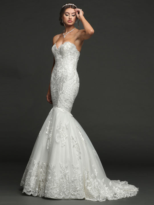 A woman models the Avery Austin TH-Julia, an ivory strapless mermaid wedding dress with lace details and a corset back, standing against a dark background while touching her forehead and wearing a necklace and headband.