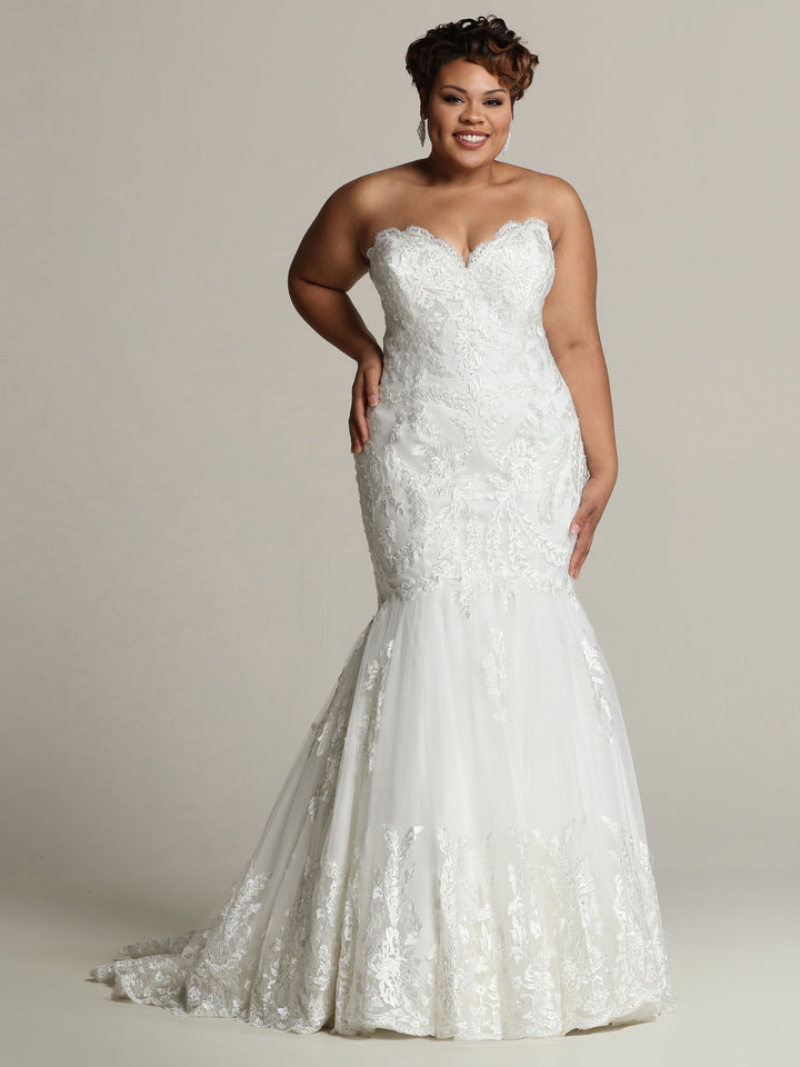 A smiling woman wears the Julia wedding dress by Avery Austin—a strapless, form-fitting lace corset gown with a sweetheart neckline and flared mermaid skirt—posing against a plain light background.