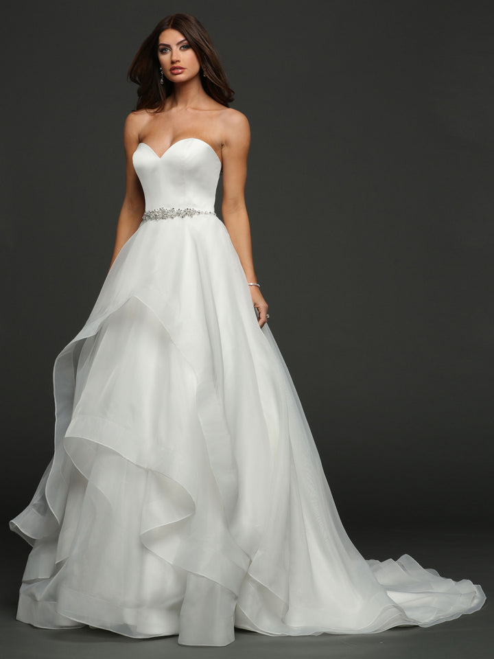 A woman in the Avery Austin "Clara" strapless ivory ball gown wedding dress with a sweetheart neckline and jeweled belt stands against a dark background. The layered fabric and slight train create an elegant bridal look.