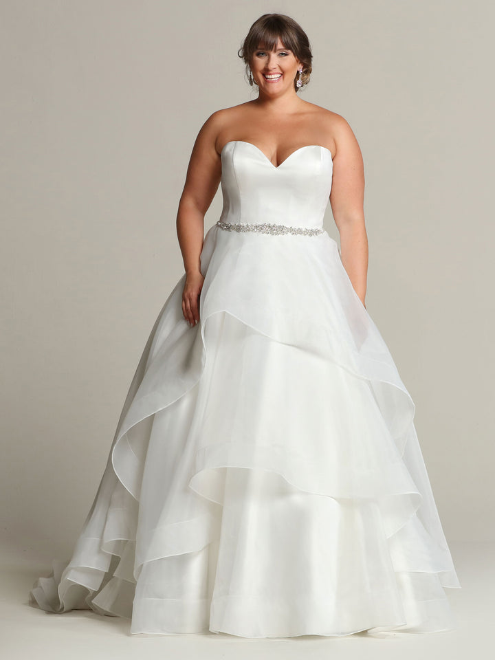 A woman smiles against a plain background wearing the Clara ball gown by Avery Austin in ivory, featuring a strapless sweetheart neckline, layered organza and satin skirt, jeweled belt, and earrings.