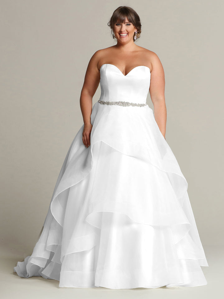 A woman in the Avery Austin "Clara" white wedding dress—strapless with a sweetheart neckline, jeweled belt, and layered satin-organza skirt—smiles against a plain background.
