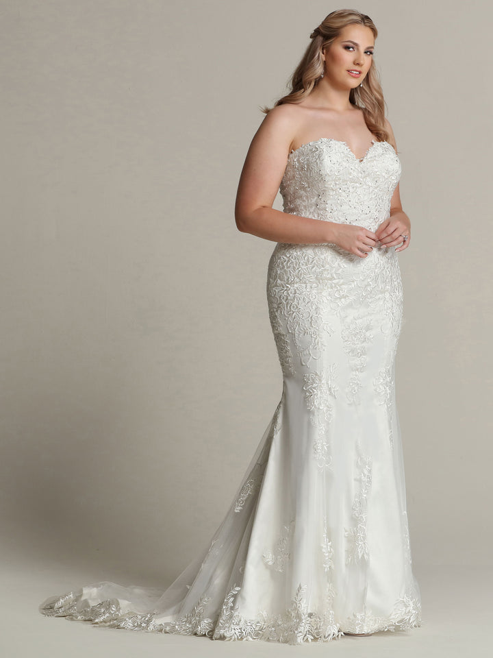 A woman with long, wavy blonde hair wears the Isla strapless wedding dress in ivory by Avery Austin, featuring delicate botanical lace appliques and a subtle train, standing against a plain light background.