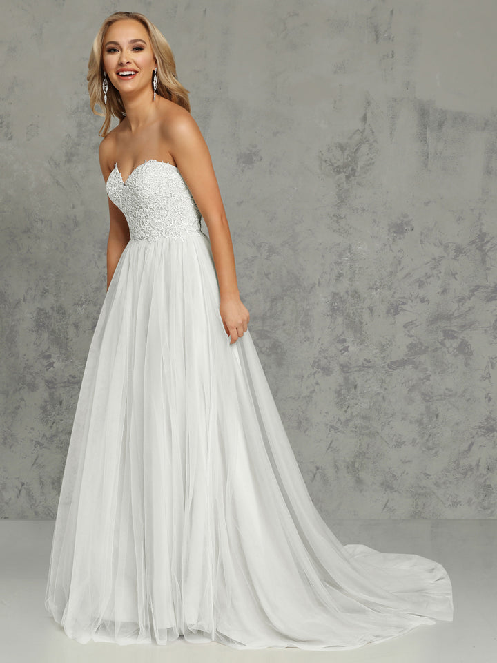 A woman smiles while wearing the Hadley wedding dress by Avery Austin in Ivory/Ivory, featuring an elegant lace bodice and flowing tulle skirt, standing against a textured gray background.