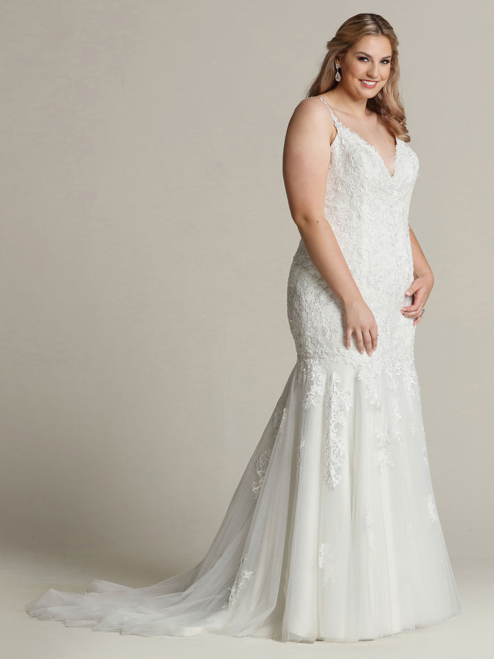 A woman smiles against a plain light background wearing the Alexandra - Tried On Sample by Avery Austin, a sleeveless ivory/nude tulle and lace mermaid wedding gown with a V-neckline and long, flowing train.
