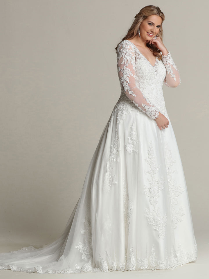 A woman models the Avery Austin "Jasmine - Tried On Sample" wedding dress in Ivory/Nude Glitter, featuring long lace sleeves, a V-neckline, and a flowing A-line skirt, smiling softly against a plain background.