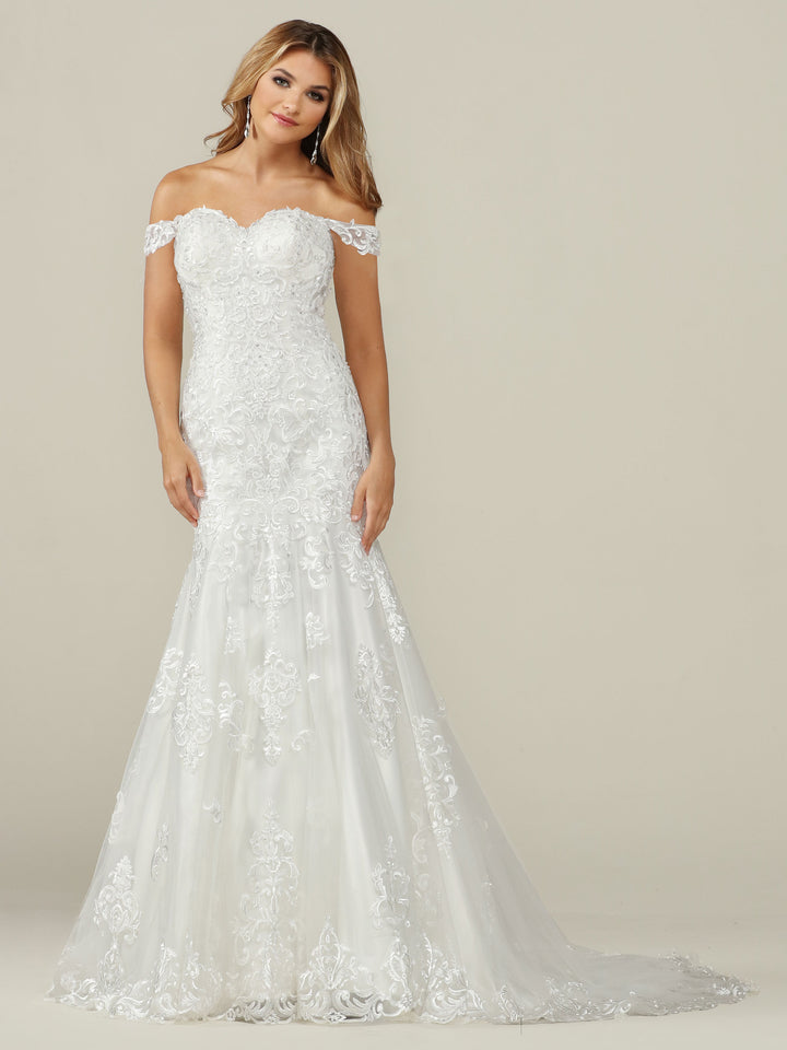 A woman in the Avery Austin TH-Juliana wedding dress, an elegant off-the-shoulder gown with a fitted bodice and flared floral lace skirt in Ivory/Ivory, stands against a plain beige background.