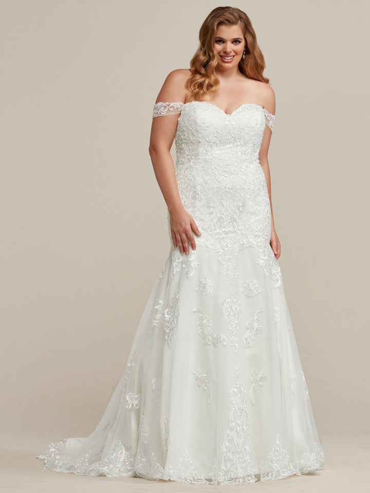 A woman smiles against a plain light background, wearing the Juliana off-the-shoulder wedding dress by Avery Austin, featuring a fitted bodice and floral lace embellishments with her hand by her side.