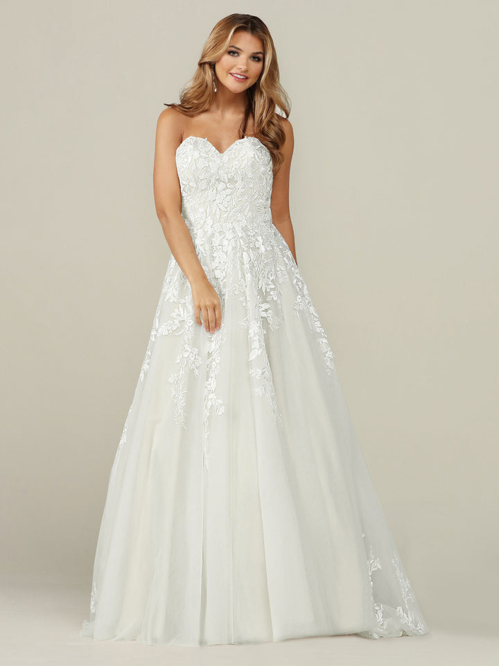 A woman smiles and looks to the side, wearing the Avery Austin TH-Emerson wedding dress in Ivory/Ivory—an elegant strapless lace gown with a sweetheart neckline, fitted bodice, and flowing A-line skirt—against a plain light background.