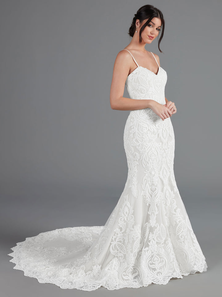 A woman models the Avery Austin TH-Morgan wedding dress in Ivory/Ivory—a lace mermaid gown with thin straps and an ornate train—standing against a gray background with hands gently clasped, showcasing its elegant design.