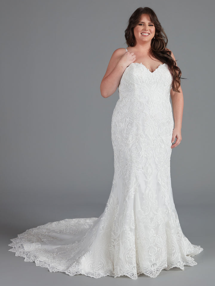 A woman with long brown hair models the Avery Austin "Morgan" strapless lace mermaid wedding dress, featuring a fitted bodice and long scalloped train, standing against a plain gray background.
