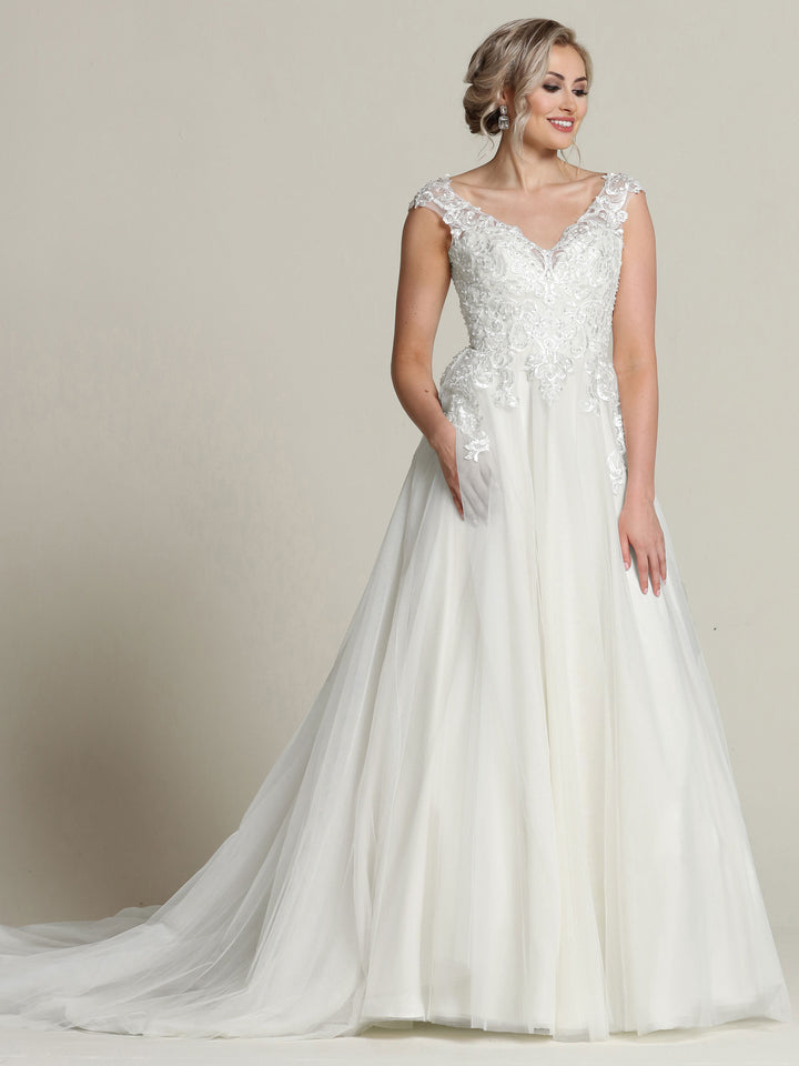 A woman in the Avery Austin "Sara" wedding dress, featuring lace cap sleeves, a delicately detailed bodice, and a flowing floor-length skirt, stands against a plain background, smiling and looking to the side.