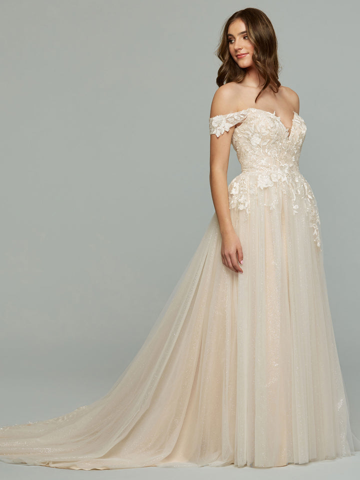 A woman in the averyaustin Trinity-G off-the-shoulder wedding dress, featuring an ivory/champagne glitter lace appliqué bodice, A-line tulle skirt, and long train, stands against a grey background, looking to the side with a gentle smile.