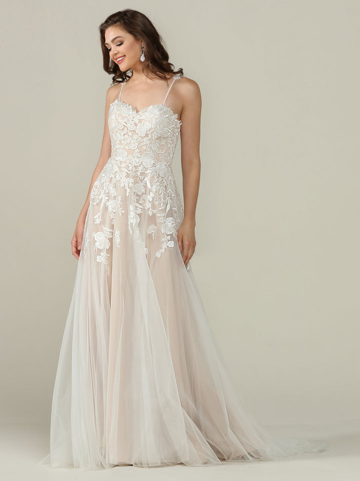 A woman in the Avery Austin TH-Esther wedding dress—sleeveless, floor-length with ivory/champagne lace, floral embroidery, and a tulle skirt—stands against a plain light background, smiling and looking to the side.