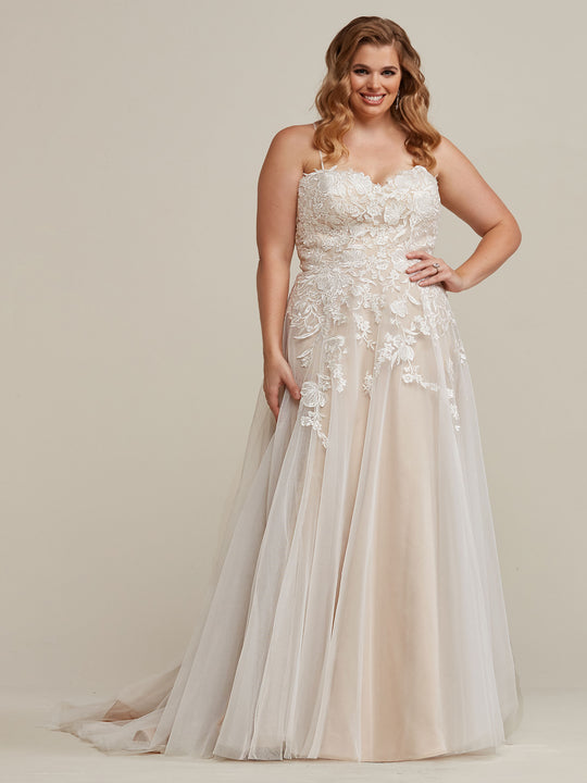 A smiling woman wears the Esther wedding dress by Avery Austin in ivory/champagne, featuring a strapless, fitted lace bodice with floral appliqués and a flowing, floor-length tulle skirt. She stands before a plain, light background.