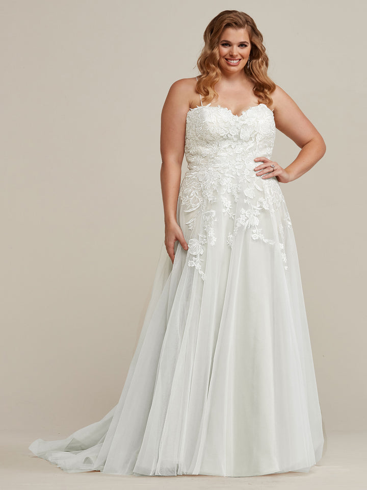 A smiling woman poses with one hand on her hip, wearing the Avery Austin TH-Esther wedding dress in Ivory/Ivory—a strapless, white lace A-line gown with a flowing tulle skirt—against a plain, light background.