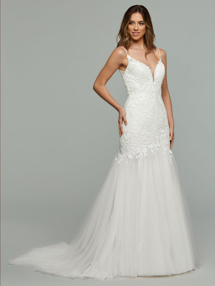 A woman models the averyaustin Imani - Tried On Sample, an ivory/ivory sleeveless mermaid wedding dress with a deep V-neckline, lace bodice, and tulle train, standing against a plain gray background.