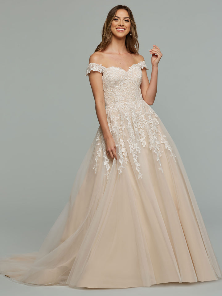A woman in the averyaustin TH-Maeve off-the-shoulder wedding dress in Ivory/Nude, with lace details and a full tulle skirt, smiles against a plain gray background.