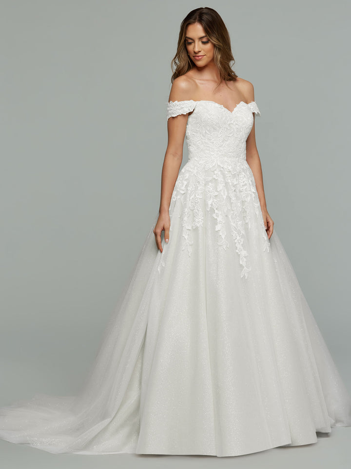 A woman wearing the averyaustin TH-Maeve off-the-shoulder lace wedding dress in Ivory/Ivory Glitter with a full tulle skirt stands against a plain gray background.
