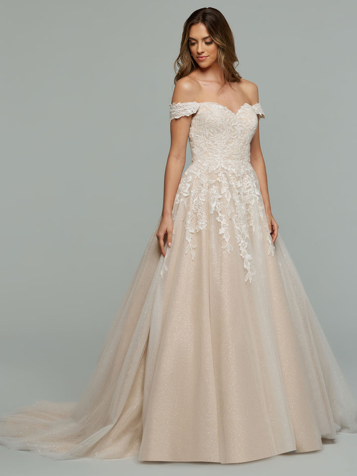 The bride wears the averyaustin Maeve-G, an off-the-shoulder lace wedding dress with delicate appliques and a glitter tulle full skirt, set against a plain gray background.