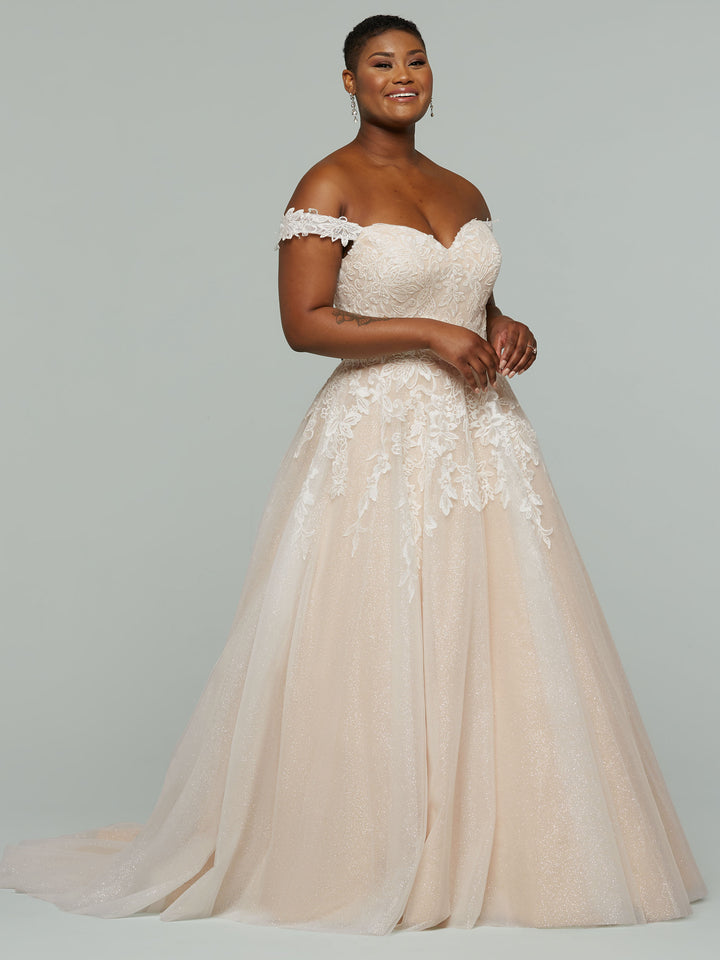 A woman with short hair and earrings smiles in the Maeve-G by averyaustin, an off-the-shoulder blush wedding dress featuring a floral lace bodice and full flowing skirt, standing against a plain background.