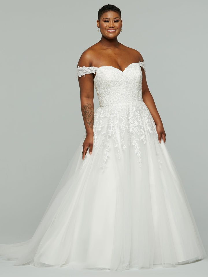 A smiling woman models the "Maeve" wedding dress by averyaustin in Ivory/Nude, featuring off-the-shoulder lace and a full tulle skirt, posing elegantly against a plain gray background.