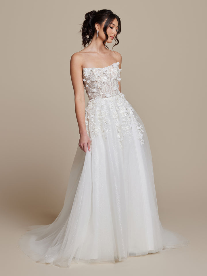 A woman in the averyaustin Opal wedding dress, featuring a strapless, sheer floral-embroidered bodice and flowing ivory/nude glitter tulle skirt, stands against a plain beige background.