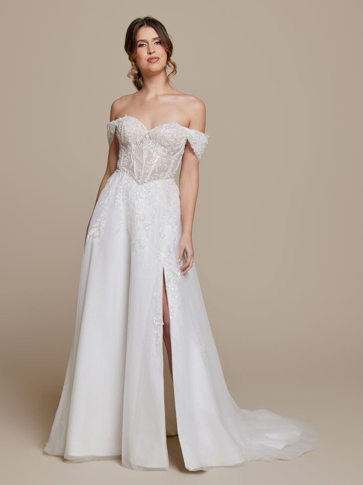 A woman wears the averyaustin Keira wedding dress in Ivory/Nude Glitter, featuring an off-the-shoulder sheer beaded lace bodice and a flowy A-line skirt with thigh-high slit, standing against a plain beige background.