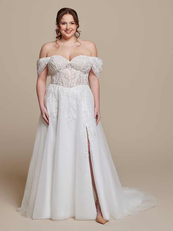 A woman wearing the Keira by averyaustin—a stunning ivory/nude glitter off-the-shoulder A-line wedding dress with beaded lace, sheer bodice, and thigh-high slit—stands and smiles against a plain beige background.