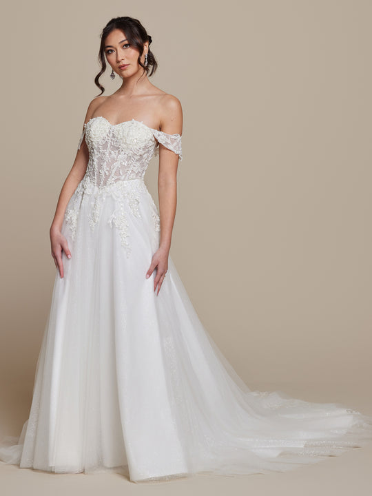 A woman in the averyaustin Daphne ivory glitter off-the-shoulder wedding dress with lace details and a long tulle train stands against a beige background, her loose, wavy hair framing her soft, elegant expression.