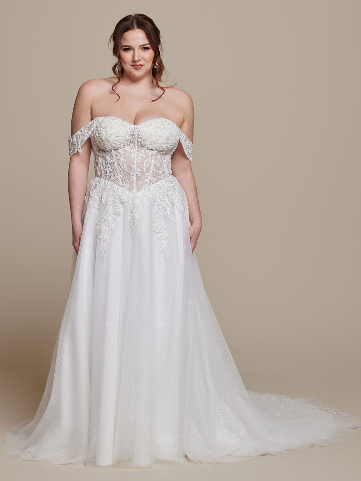 A woman wearing the Daphne wedding dress by averyaustin in Ivory Glitter, featuring an off-the-shoulder design, sheer lace bodice, and flowing A-line skirt, stands against a plain beige background.