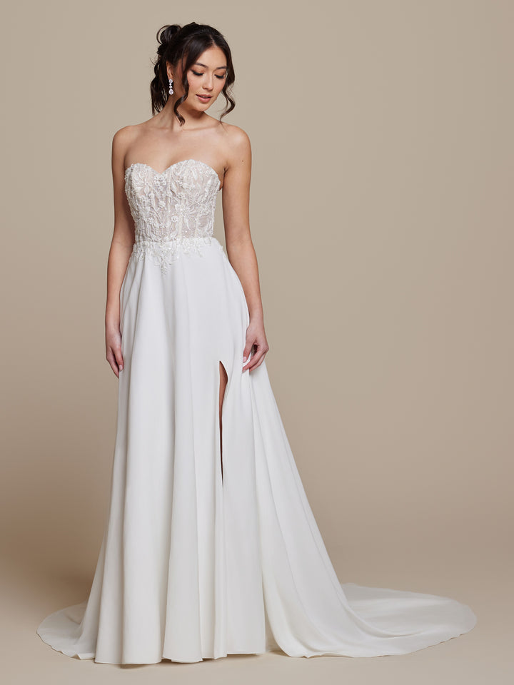 A woman wearing the Aurielle strapless tulle corset wedding dress by averyaustin in Ivory/Nude Glitter, featuring a lace-embellished bodice, flowing A-line skirt with thigh-high slit, stands against a plain beige background.