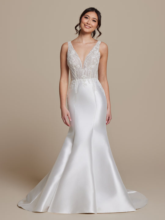A woman smiles against a beige background wearing the Colette by averyaustin, a sleeveless fitted beaded lace wedding dress in Ivory/Nude Glitter with a deep V-neckline, flared satin skirt, and long train in classic fit-and-flare style.