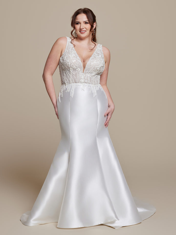 A woman smiles against a beige background, wearing "Colette" by averyaustin—an ivory/nude glitter, sleeveless beaded lace wedding dress with a deep V-neckline and fit-and-flare silhouette.